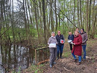 Sechs Menschen stehen in einer Gruppe vor einem Wald. Der Waldboden ist nass.