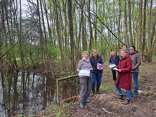 Sechs Menschen stehen in einer Gruppe vor einem Wald. Der Waldboden ist nass.