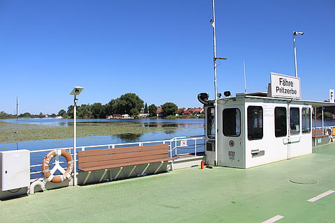 Autofähre in Pritzerbe mit grünem Belag und weißem Fährhaus. Dahinter blaues Wasser der Havel mit Seerosenfeld.