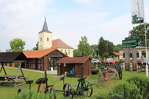 Alte landwirtschaftliche Geräte stehen auf einer grünen Wiese. Im Hintergrund sieht man den Gebäude und den Kirchturm von Großderschau.