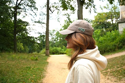 Junge Frau mit langen Haaren, weißem Kapuzenpullover und einer Schirmmütze mit einem Brandenburg anstecker steht im Profil auf einem Waldweg. 
