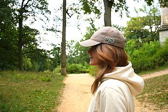 Junge Frau mit langen Haaren, weißem Kapuzenpullover und einer Schirmmütze mit einem Brandenburg anstecker steht im Profil auf einem Waldweg. 