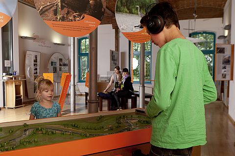Zwei Kinder stehen mit Kopfhörern vor einem Wassermodell.